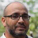 A man with glasses, a beard, and a shaved head is outdoors in a blurred green setting. He is wearing a light-colored shirt. .